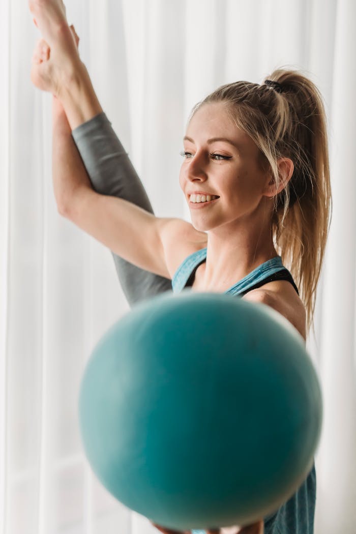 Positive young female athlete with raised leg holding ball while training against white curtain on window
